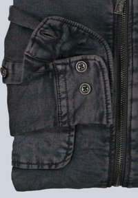 Schwarze gefaltete Jacke mit geknöpftem Klappenfach, Reißverschluss und strukturiertem Stoffdetail auf hellem Hintergrund.