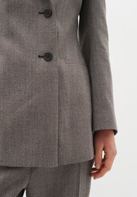 Šedý herringbone blazer se třemi černými knoflíky, klopovým límcem, texturovanou tkaninou a bočními kapsami, spárovaný se shodnými kalhotami.