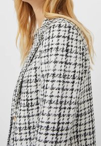 White and black tweed blazer with a check pattern, featuring a notched collar and a button closure, showing textured fabric and structured design.