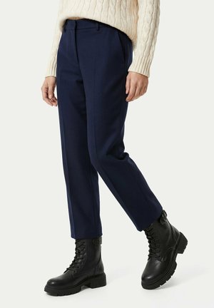 Person trägt marineblaue, maßgeschneiderte Hose, cremefarbenen Kabelstrickpullover und schwarze Schnürstiefeletten mit dicken Sohlen.