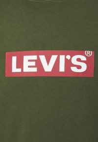 Olivgrön t-shirt med en framträdande röd rektangulär grafik med vit "LEVI'S®" text i fet stil. Slät textur, avslappnad design.