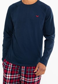 Marineblaues Langarmshirt mit Rundhalsausschnitt und rotem Akzentlogo. Kombiniert mit rot-schwarz karierter Schlafanzughose mit elastischem Bund.