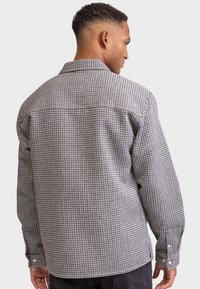 Ein Mann, der mit dem Rücken zur Kamera steht, trägt ein graues gemustertes Langarmhemd mit geknöpften Manschetten und schwarze Hose vor einem einfarbigen Hintergrund.