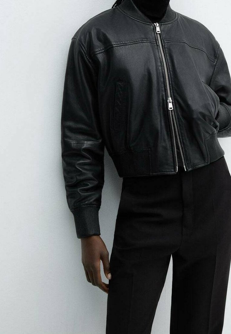 Personne portant une veste bomber en cuir noire zippée et un pantalon noir taille haute, debout devant un fond gris clair uni.