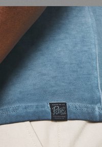 Blaues Baumwoll-T-Shirt mit weicher Textur. Mit einem schwarzen Etikett "CRUDE OIL CO", das am Saum angenäht ist. Leicht verblasste Farbe, lässiges Design.