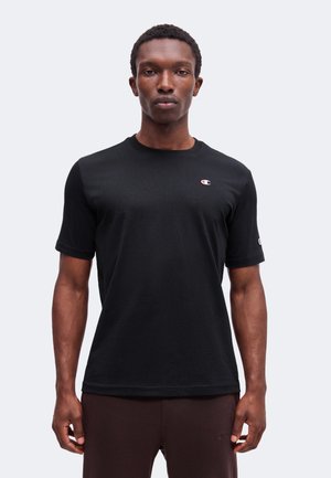 Jeune homme de face portant un T-shirt noir à manches courtes avec un petit logo Champion sur la poitrine et la manche, debout devant un fond uni.