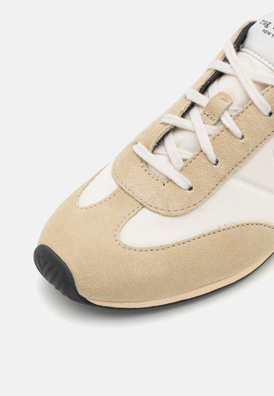 Beige suede sneaker featuring a smooth leather upper, flat laces, rounded toe, and a black rubber sole with subtle stitching detail.