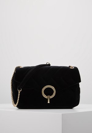 Sac à bandoulière en velours matelassé noir avec fermeture circulaire dorée et anse chaîne, posé sur une surface blanche.