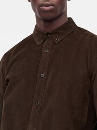 Homme portant une chemise en velours côtelé marron foncé boutonnée avec un col boutonné sur un fond uni clair.