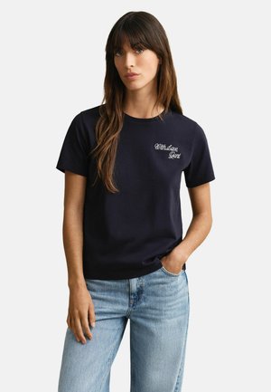 Frau mit langem braunem Haar, die ein schwarzes T-Shirt mit dem Schriftzug "With Love, Gant" und hellblaue Jeans trägt, steht mit einer Hand in der Tasche.