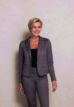 Smiling woman with short blonde hair wearing a matching gray blazer and pants over a black top, standing against a plain light wall.