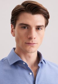 Jonge man met bruin haar in een blauw patroonhemd, kijkt recht vooruit met een neutrale uitdrukking op een lichte achtergrond.