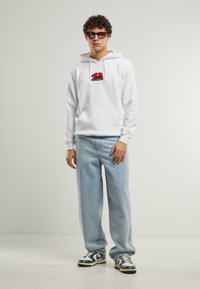 Vit hoodie med röd björngrafik och texten "California", i kombination med ljusblå baggy jeans och gröna och vita sneakers.