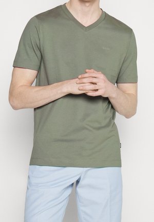 Mann trägt ein olivgrünes V-Ausschnitt-T-Shirt mit kurzen Ärmeln und "BOSS"-Logo sowie hellblaue Hosen, die Hände vor dem Körper verschränkt.