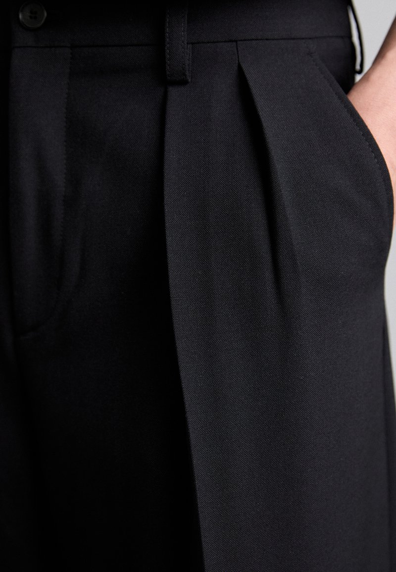 Black pleated trousers made of smooth fabric, featuring a waistband, side pockets, and a button detail on the waist.
