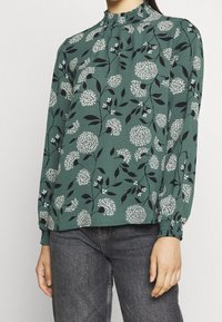 Grön blommig blus med lång ärm, hög rynkad hals, elastiska manschetter och mönster av vita blommor och svarta blad.