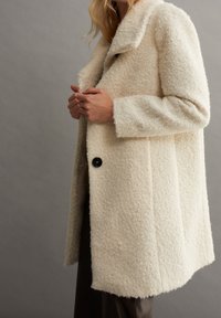 Crèmekleurige jas van textuurstof, met een grote kraag, enkele knoopsluiting en brede mouwen. Eenvoudig ontwerp.