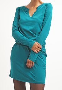 Teal pitkähihaine mekko, jossa on V-puoli ja nappidetalji. Sileä kangas, kiristetty vyötärö ja rento istuvuus, ulottuen reiteen puoliväliin.