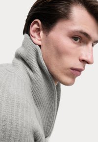 Suéter de punto acanalado gris con un cuello alto, que presenta una textura suave y costuras prominentes, llevado por un modelo visto de perfil.