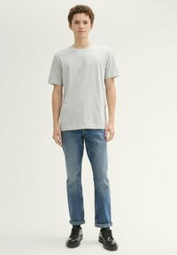 Joven de pie en una pose neutral, vistiendo una camiseta gris claro, vaqueros azules remangados y zapatos de vestir negros, sobre un fondo liso.