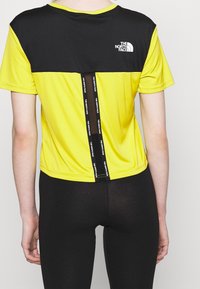 Żółta i czarna koszulka sportowa z krótkimi rękawami, z logo na klatce piersiowej oraz czarnym akcentem z tyłu, z detalami marki.