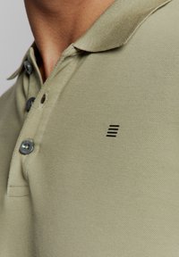 Olivengroene polo-shirt van textuurstof, met een drieknops revers en een klein zwart logo op de borst.