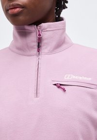 Κοντινό πλάνο ενός ατόμου που φοράει ένα ανοιχτό μωβ φλις πουλόβερ με μισό φερμουάρ και τσεπάκι με φερμουάρ στη δεξιά πλευρά του στήθους.