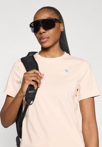 Lyserød t-shirt med korte ærmer og et blåt trekantet logo, iført sorte oversized solbriller og holder fast i stroppen til en sort rygsæk.