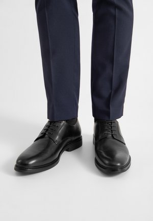 Mustad musta nahast ujumisjalanõusid ümarate varvastega, sileda tekstuuriga ja sünteetiliste paeltega, kombineeritud tumesiniste kitsaste pükstega.