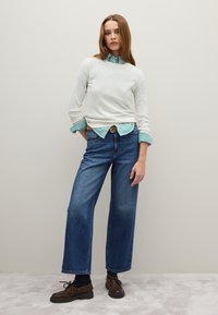 Weißer gerippter Pullover über einem grün-weiß gestreifen Hemd, kombiniert mit weit geschnittenen blauen Jeans und braunen Schnürschuhen.