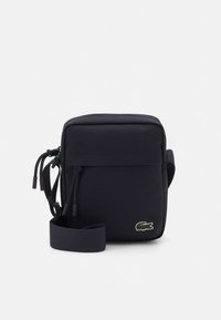 Lille sort crossbodytaske med lynlåslomme foran, justerbar rem og grønt krokodillelogo i nederste højre hjørne.