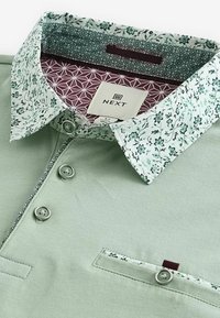 Polo vert clair avec col et bordure de poche à motif floral, trois boutons et doublure intérieure du col à motif géométrique.