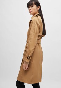 Beige Trenchcoat aus glattem Material mit tailliertem Design, Knopfleiste und verstellbaren Ärmelbündchen. Das Modell steht im Profil.