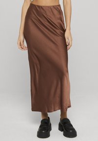 Jupe longue en satin marron à la texture lisse, taille élastique, et silhouette droite. Portée avec des mocassins noirs à semelle épaisse.