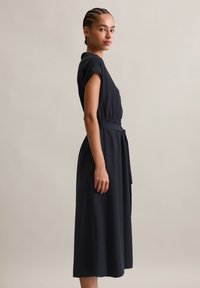 Junge Frau mit geflochtenem Haar, die ein ärmelloses schwarzes Midikleid mit gebundenem Taillengürtel trägt, steht im Profil vor einem einfarbigen Hintergrund.