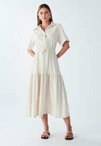 Šviesiai ruda, vidutinio ilgio suknelė trumpomis rankovėmis, su sagų priekiu, elastingu juosmeniu ir sluoksniuotu sijonu. Lygus, tekstūruotas audinys su kasdiene dizaino koncepcija.