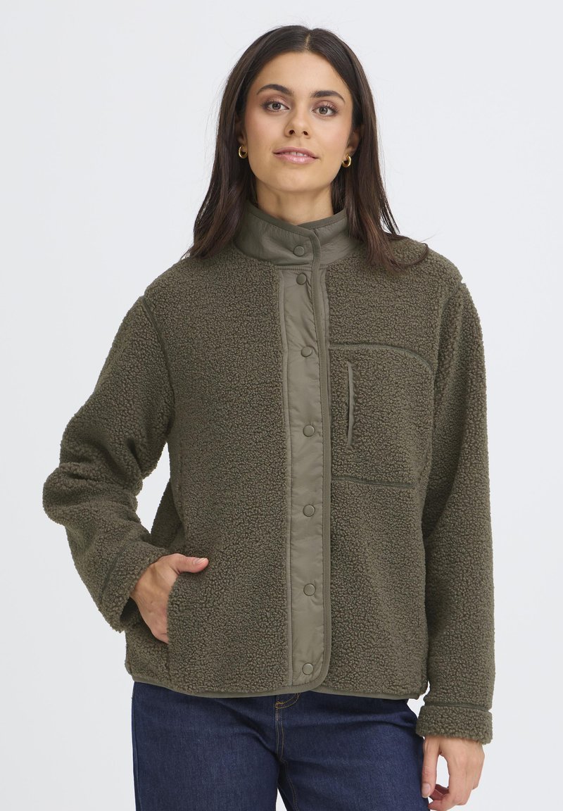 Kvinde iført olivengrøn fleecejakke med trykknapper, der står med den ene hånd i lommen mod en ensfarvet baggrund.