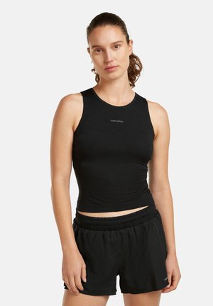 Mujer con camiseta deportiva negra sin mangas y pantalones cortos negros, de pie contra un fondo claro y liso.