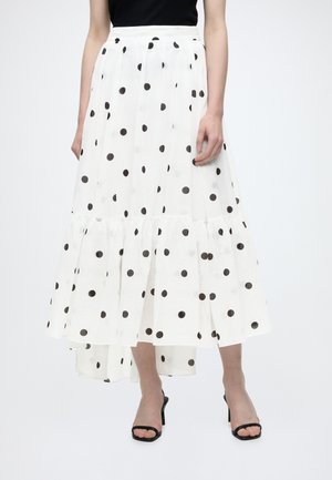 Femme portant une jupe blanche taille haute, longueur cheville, à pois noirs, un haut noir et des talons noirs ouverts, debout droite.