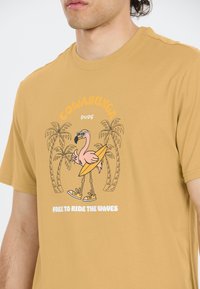 Beige bomulls t-shirt med en flamingo som har solglasögon och en surfbräda, omgiven av palmer, med texten "COWABUNGA DUDE" ovanför.