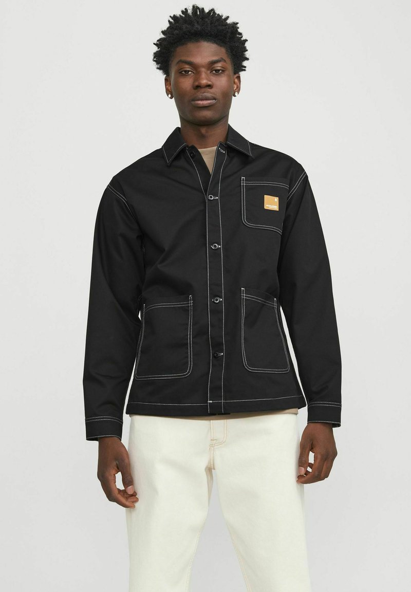 Black, long-sleeve jacket with white stitching, front button closure, chest pocket, and two front pockets; worn over light-colored pants.