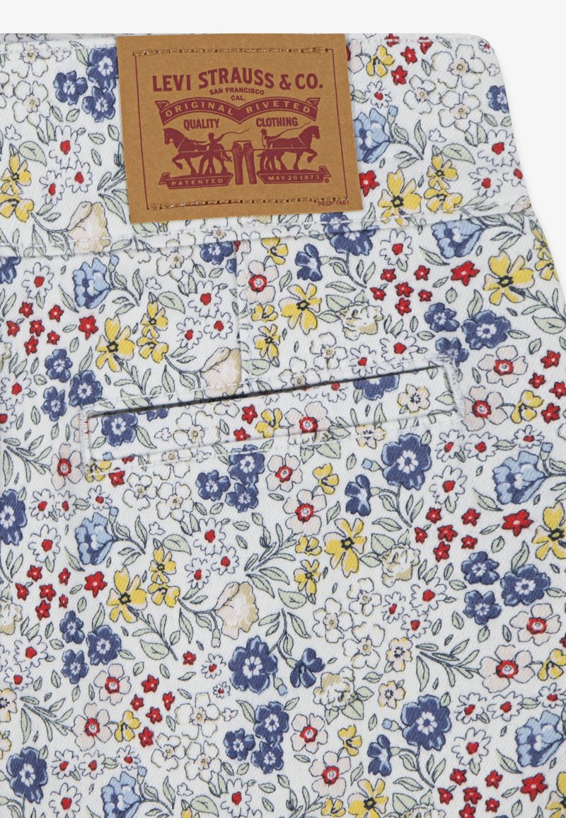 Tissu à motif floral en blanc avec des fleurs bleues, rouges et jaunes. Présente une étiquette marron avec du texte embossé et des détails de design.