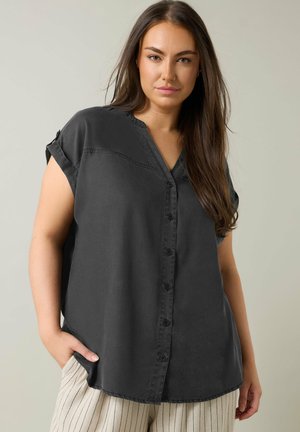 Kobieta z długimi brązowymi włosami, ubrana w luźną czarną koszulę bez rękawów zapinaną na guziki oraz jasne spodnie w paski, stojąca z jedną ręką w kieszeni.