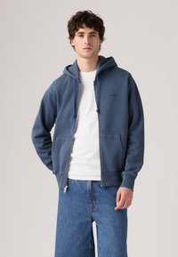 Ung mann iført en blå hettejakke med glidelås over en hvit skjorte og blå jeans, stående mot en enkel lys bakgrunn.