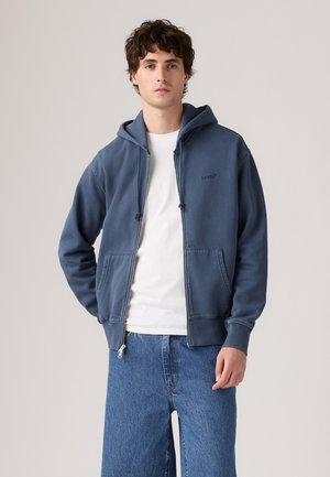 Jonge man in een blauwe hoodie met rits over een wit shirt en blauwe spijkerbroek, staand tegen een effen lichte achtergrond.