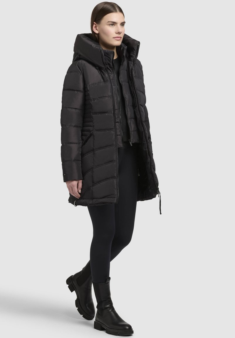 Manteau doudoune noir avec un col montant, doté d'une fermeture éclair à l'avant et d'un design matelassé. Porté sur des leggings noirs et des bottes noires épaisses.