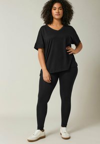 Zwarte oversized V-hals t-shirt met opgerolde mouwen, gecombineerd met zwarte leggings en witte sneakers met tan kleurige zolen. Glad stof, ontspannen pasvorm.