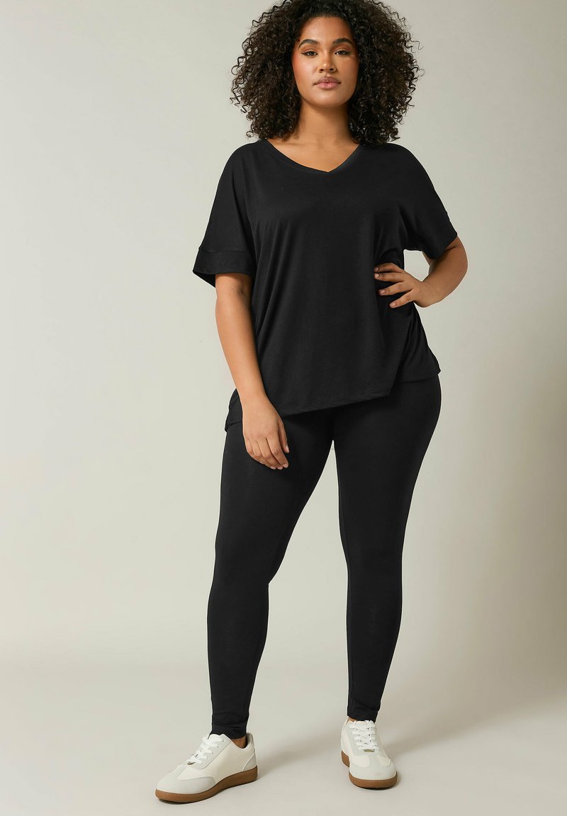 Zwarte oversized V-hals t-shirt met opgerolde mouwen, gecombineerd met zwarte leggings en witte sneakers met tan kleurige zolen. Glad stof, ontspannen pasvorm.