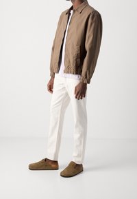 Brun lättviktig jacka med elastiska manschetter, tillsammans med en vit T-shirt och vita byxor. Bruna glidklädda träskor har en spänne detalj.