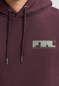 Bordeauxfarbener Kapuzenpullover mit Kordelzug, ausgestattet mit einem reflektierenden Logo auf der Brust und silbernen Enden an den Kordeln. Weiche Stoffoberfläche.